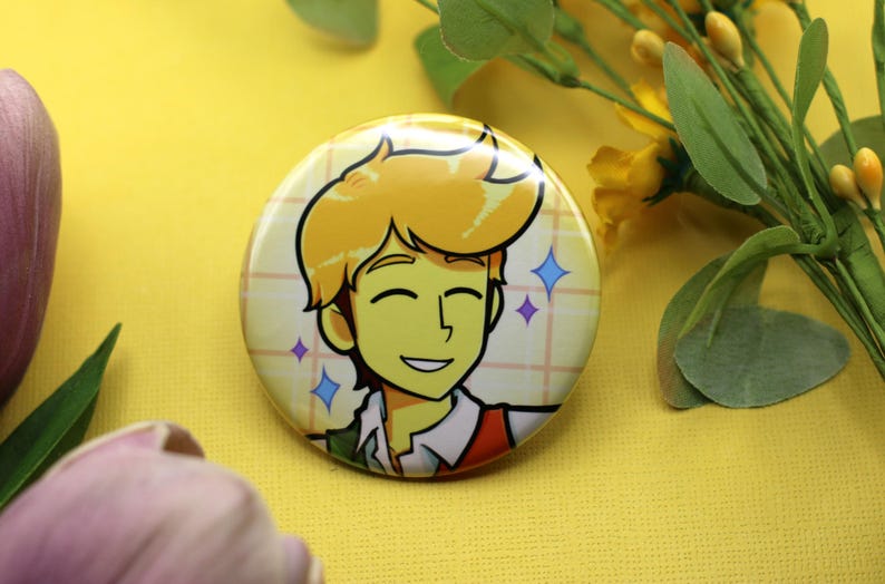 K&ouml;nnte beinhalten: Ein runder Ansteckbutton mit einer Cartoon-Illustration einer l&auml;chelnden Person mit blonden Haaren, die ein wei&szlig;es Hemd mit Kragen und eine rote Weste tr&auml;gt. Der Hintergrund der Illustration ist ein hellgelbes Gittermuster mit kleinen Sternsymbolen.