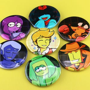 K&ouml;nnte beinhalten: Eine Sammlung von sieben runden Ansteckbuttons mit farbenfrohen Cartoon-Charakter-Illustrationen. Die Motive zeigen eine Figur mit violettem Haar, eine Person mit blauem Haar, eine gr&uuml;nh&auml;utige Figur, eine gelbhaarige Person und ein orangefarbenes Wesen.