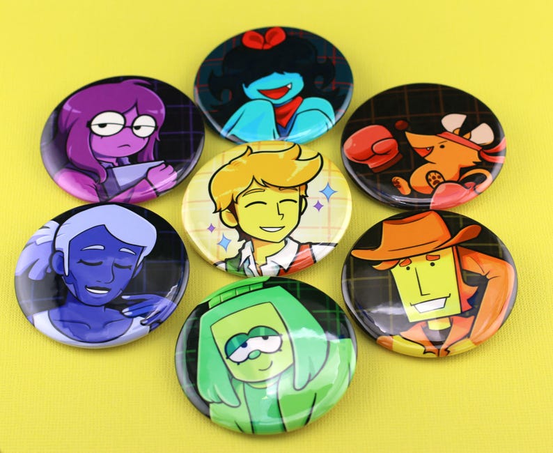 K&ouml;nnte beinhalten: Eine Sammlung von sieben runden Ansteckbuttons mit farbenfrohen Cartoon-Charakter-Illustrationen. Die Motive zeigen eine Figur mit violettem Haar, eine Person mit blauem Haar, eine gr&uuml;nh&auml;utige Figur, eine gelbhaarige Person und ein orangefarbenes Wesen.