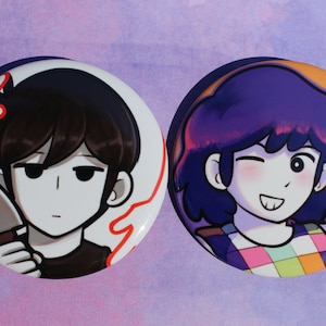 May include: Two round button badges featuring cartoon characters. The first badge features a character with black hair holding a knife. The second badge features a character with purple hair and a colourful checked shirt.