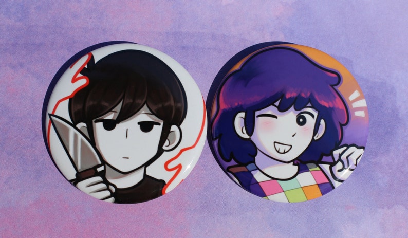 May include: Two round button badges featuring cartoon characters. The first badge features a character with black hair holding a knife. The second badge features a character with purple hair and a colourful checked shirt.