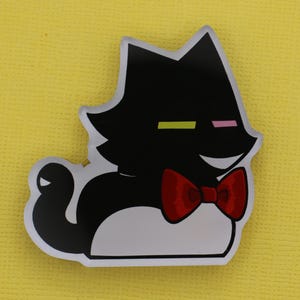 Puede incluir: Una pegatina con forma de gato negro con pajarita roja. El gato tiene la barriga blanca y una cara sonriente. Los ojos del gato est&aacute;n representados por una l&iacute;nea horizontal amarilla y rosa. La pegatina est&aacute; sobre un fondo amarillo.
