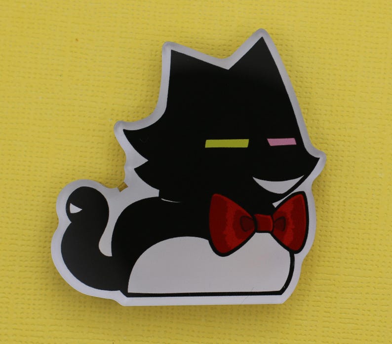 Puede incluir: Una pegatina con forma de gato negro con pajarita roja. El gato tiene la barriga blanca y una cara sonriente. Los ojos del gato est&aacute;n representados por una l&iacute;nea horizontal amarilla y rosa. La pegatina est&aacute; sobre un fondo amarillo.