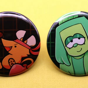 K&ouml;nnte beinhalten: Zwei runde Ansteckbuttons. Der linke Button zeigt eine orangefarbene Maus mit rotem Stirnband und rotem Boxhandschuh vor einem dunklen Gitterhintergrund. Der rechte Button zeigt eine gr&uuml;ne Zeichentrickfigur mit Kapuze vor einem dunklen Gitter.
