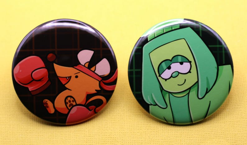 K&ouml;nnte beinhalten: Zwei runde Ansteckbuttons. Der linke Button zeigt eine orangefarbene Maus mit rotem Stirnband und rotem Boxhandschuh vor einem dunklen Gitterhintergrund. Der rechte Button zeigt eine gr&uuml;ne Zeichentrickfigur mit Kapuze vor einem dunklen Gitter.