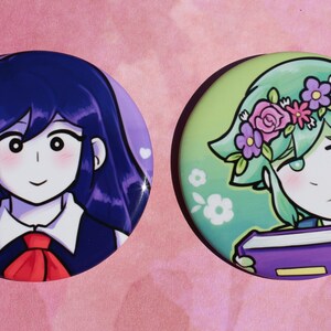 May include: Two round button badges featuring anime-style characters. The first badge features a girl with long, dark blue hair, wearing a white collared shirt with a red bow tie. The second badge features a girl with short, green hair wearing a flower crown and holding a purple book.