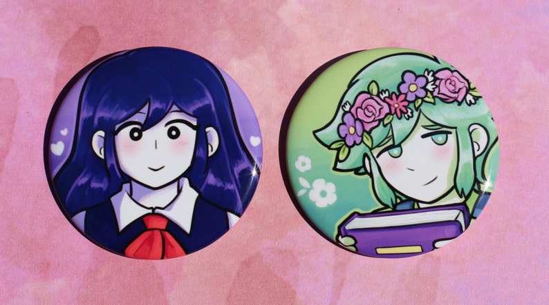 May include: Two round button badges featuring anime-style characters. The first badge features a girl with long, dark blue hair, wearing a white collared shirt with a red bow tie. The second badge features a girl with short, green hair wearing a flower crown and holding a purple book.