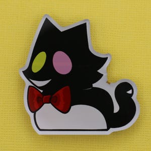 Puede incluir: Un personaje de dibujos animados en blanco y negro con forma de gato, con pajarita roja, ojos verdes y rosas y una sonrisa. Sobre un fondo amarillo.