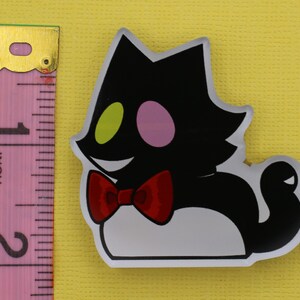 Puede incluir: Una pegatina con forma de gato negro con pajarita roja, ojos verdes y rosas y vientre blanco. La pegatina mide aproximadamente 5 cm de alto, medido con la regla rosa.