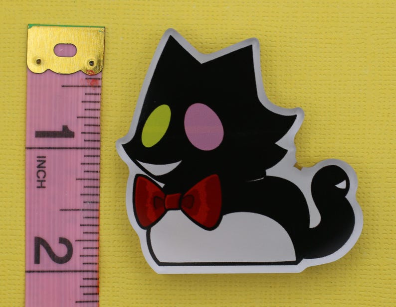 Puede incluir: Una pegatina con forma de gato negro con pajarita roja, ojos verdes y rosas y vientre blanco. La pegatina mide aproximadamente 5 cm de alto, medido con la regla rosa.