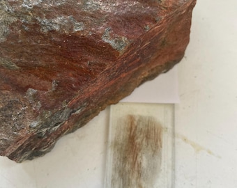 May include: A large, rough-textured rock with reddish-brown and gray hues. A clear microscope slide with a sample of the rock is positioned below the rock, showing a detailed view of the rock's composition.