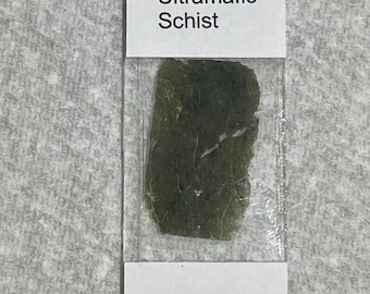 May include: A dark green Ultramafic Schist rock sample encased in a clear plastic slide. The sample is rectangular with a rough texture. The text "Ultramafic Schist" is printed on a white label above the sample.