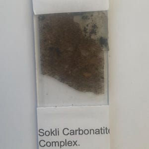 Può includere: Vetrino microscopico con un campione del Complesso di Carbonatite di Sokli, Finlandia. Il campione è un mix di materiale granulare marrone e nero, parzialmente ricoperto da un adesivo trasparente. Il vetrino ha un'etichetta bianca.