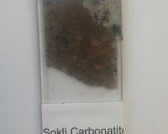 May include: A microscopic slide with a sample of Sokli Carbonatite Complex from Finland. The sample is a mix of brown and black granular material, partially covered by a clear adhesive. The slide has a white label with the sample's name and origin.