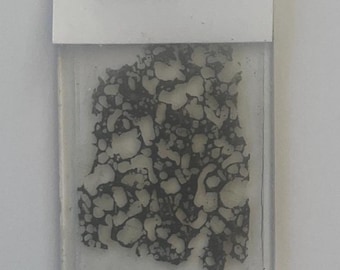 May include: A geological sample of Oceanite-Vesicular Porphyritic Olivine Basalt from Hawaii, USA. The sample is encased in a clear rectangular holder. The rock has a dark gray and white speckled pattern.