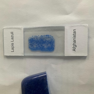 Peut inclure: Un échantillon de Lapis Lazuli d'Afghanistan, présenté sur une lame de microscope. La lame montre un échantillon bleu et granuleux. Une pierre de Lapis Lazuli polie, bleu foncé, est également présente, mettant en valeur sa couleur et sa texture profondes.