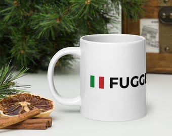 May include: White ceramic mug with the word "FUGGE" in black letters. The letter "F" is preceded by the green and red stripes of the Italian flag. The mug is on a white surface with a dried orange slice and cinnamon sticks.