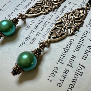 May include: A pair of dangle earrings featuring iridescent green pearl beads and ornate bronze-colored filigree accents. The earrings have a vintage aesthetic, with detailed floral and scrollwork designs.