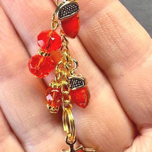 May include: A gold-toned charm featuring red acorn and maple leaf pendants, along with red faceted beads. The acorn caps have a silver-colored design. The charm is designed for jewelry or accessories.