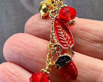 May include: A gold charm with autumnal pendants. It features red crystal beads, a red leaf, and red acorns with black caps. The charm is suspended from a gold ring.