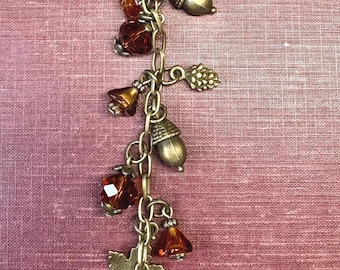 May include: A bronze-toned metal keyring featuring a vertical chain adorned with various charms including a squirrel, maple leaves, pinecones, acorns, and faceted amber-coloured glass beads attached with small metal jump rings and a lobster clasp.