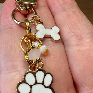 May include: Gold-tone keychain with a dog-themed design. It features a white paw print charm, a white bone charm, and small white and pink beads. The keychain has a lobster clasp for easy attachment.