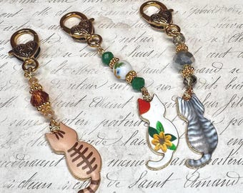 May include: Three charm necklaces with gold lobster clasps and cat charms. The charms are painted in different colors and patterns, including white with red and yellow accents, gray and white stripes, and brown and white stripes.