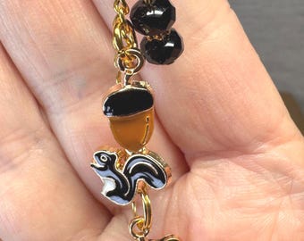 May include: A gold-coloured keyring with a charm featuring a black and white squirrel, an acorn, a leaf, and two black beads. The squirrel has a white belly and a black tail. The acorn is brown with a black top.