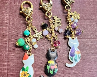 May include: Three gold keychains with cat charms. Each charm features a different cat design with floral accents. The charms are adorned with colorful beads and charms, including butterflies, flowers, and crystals.