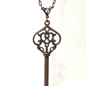 May include: A bronze key pendant necklace featuring a decorative, ornate key design. The key has a long, slender shaft and a detailed, intricate head.