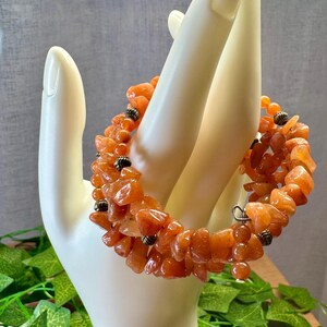 May include: A multi-strand bracelet with orange, irregularly shaped beads. The bracelet is accented with small, dark metal beads and is displayed on a white hand-shaped display. The background is a mix of grey and green.