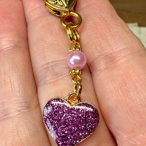 May include: A gold-colored heart-shaped clasp with a dangling charm. The charm features a purple glitter heart pendant, a pink bead, and gold-colored chain links. The clasp has a heart design.