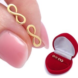 May include: A pair of gold infinity symbol earrings in a red heart-shaped box. The earrings are small and delicate.