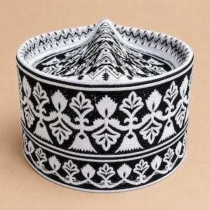Könnte beinhalten: Eine strukturierte zylindrische Kopfbedeckung mit schwarzer Basis und aufwendiger weißer Stickerei. Das Design umfasst wiederkehrende florale Motive an den Seiten und ein geometrisches Zickzackmuster am oberen Rand, abgerundet durch eine weiße Paspelierung.