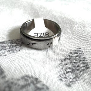 May include: A silver-colored spinner ring with etched symbols and the word "SIZE" on a white tag. The ring features a lightning bolt and other small symbols. The ring is on a white patterned surface.
