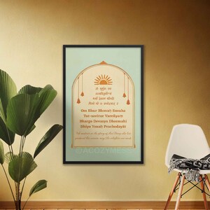May include: A framed print with a light blue background and gold accents. The print features a Sanskrit mantra in gold lettering, with a sun and bells at the top. The text reads "Om Bhur Bhuvah Suvaha Tat-savitur Varenyam Bhargo Devasya Dheemahi Dhiyo Yonah Prachodayat". The print also includes the text "We meditate on the glory of that Being who has produced the universe, may she enlighten our minds."