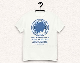 Puede incluir: Camiseta blanca con un diseño circular azul que presenta una silueta de una persona mirando las estrellas. El texto dice "Dwell on the beauty of life. Watch the stars, and see yourself running with them. - Marcus Aurelius"