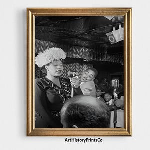 Puede incluir: Impresión enmarcada en blanco y negro que representa a una cantante de jazz actuando en el escenario. La cantante lleva un vestido oscuro y un sombrero con plumas, sosteniendo un micrófono. Hay otras personas en el fondo. El marco es dorado.