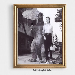 May include: Framed black and white print featuring a woman standing next to a Godzilla statue holding an umbrella. The woman is wearing a white shirt, black pants, and white shoes. The print is in a gold frame. Text at the bottom reads "ArtHistoryPrintsCo."
