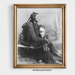 Puede incluir: Impresión enmarcada en blanco y negro de un hombre nativo americano sentado, con ropa tradicional y un tocado de plumas. El retrato está sobre un fondo neutro, con una planta en maceta visible. El marco es dorado.