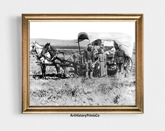 May include: A framed black and white photograph depicts a horse-drawn covered wagon with a group of people standing beside it. The horses are hitched to the wagon, and the people are dressed in period clothing. The photograph is set against a backdrop of a grassy field and a distant horizon.