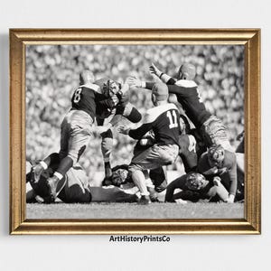 Puede incluir: Impresión enmarcada en blanco y negro de un partido de fútbol americano vintage. Los jugadores con cascos y uniformes están en acción, con una multitud al fondo. El marco es dorado.