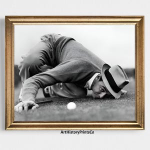 May include: A black and white photograph of a man in a hat looking at a golf ball on the green.