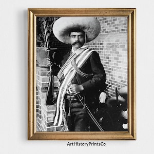May include: Framed black and white print of a man in traditional Mexican clothing. He wears a large sombrero, a bandolier, and holds a rifle. The print is in a gold frame and is labelled "ArtHistoryPrintsCo".