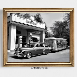 May include: Black and white framed print of a vintage scene at a Shell gas station. A classic car is parked in front of the station, with a travel trailer hitched behind it. The Shell logo is visible.