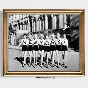 May include: A framed black and white photograph featuring a group of women in matching outfits spelling out "NEW YORK". They wear black shorts, white tops, and bow ties. The image is set against a backdrop of a building and a sidewalk.