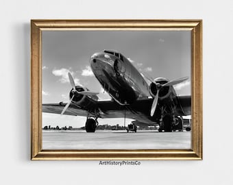 Op de afbeelding: Zwart-wit ingelijste prent van een vintage vliegtuig op een landingsbaan. Het vliegtuig heeft twee propellers en staat tegen een bewolkte lucht. De prent zit in een goudkleurige lijst.