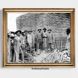 May include: A framed black and white photograph featuring a group of men in hats and suits standing in front of a brick wall. Rifles are stacked in the foreground. The photograph is in a gold-colored frame.