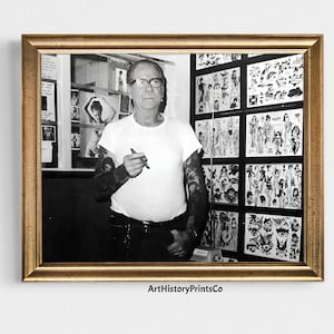 Könnte beinhalten: Schwarzweißfoto eines tätowierten Mannes, der in einem Tattoo-Studio steht. Er trägt ein weißes T-Shirt und hält einen Stift in der Hand. Die Wände sind mit gerahmten Tattoo-Designs ausgekleidet.