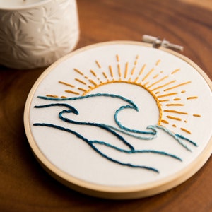 Peut inclure: Art de broderie en forme de cercle repr&eacute;sentant une vague bleue avec un soleil jaune qui se l&egrave;ve au-dessus.